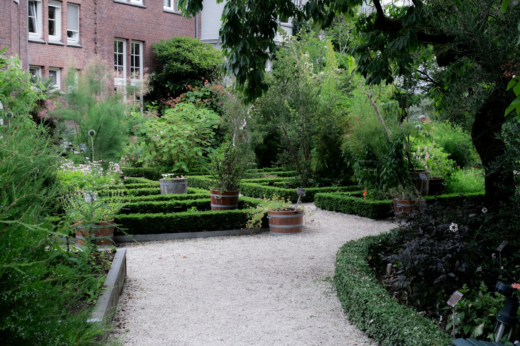A landscaped garden with clipped hedges, gravel paths, potted plants, mature trees, and brick buildings beyond.
