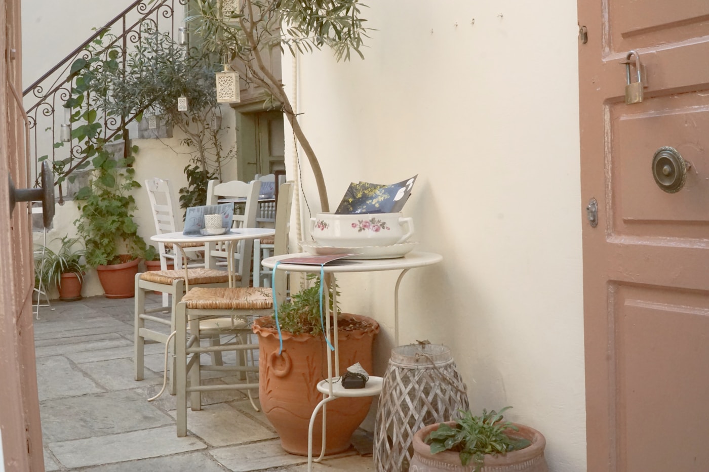 A small stone patio furnished with tables and chairs, surrounded by potted plants and an olive tree.